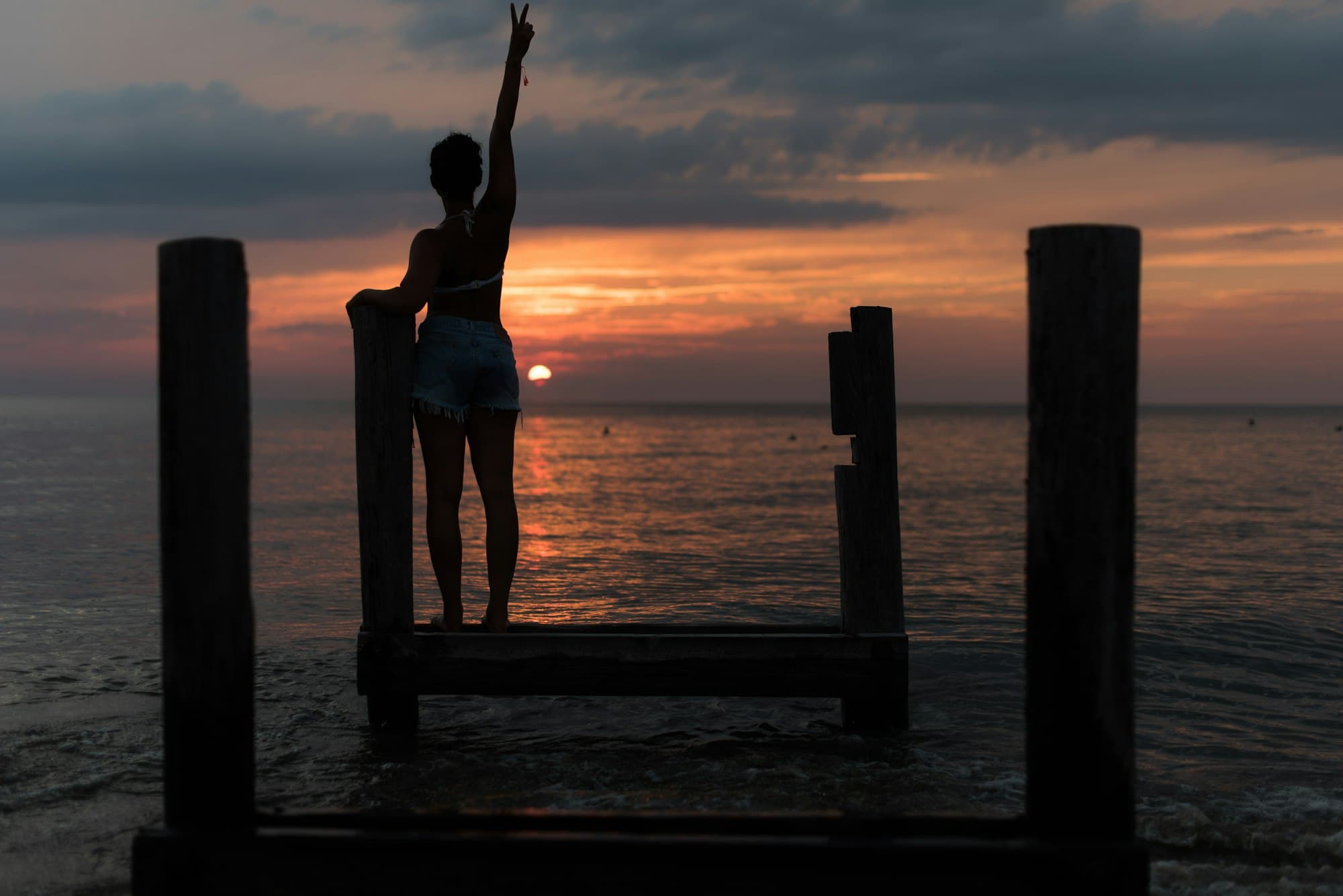 Person on a wooden dock raising an arm toward the setting sun over the water